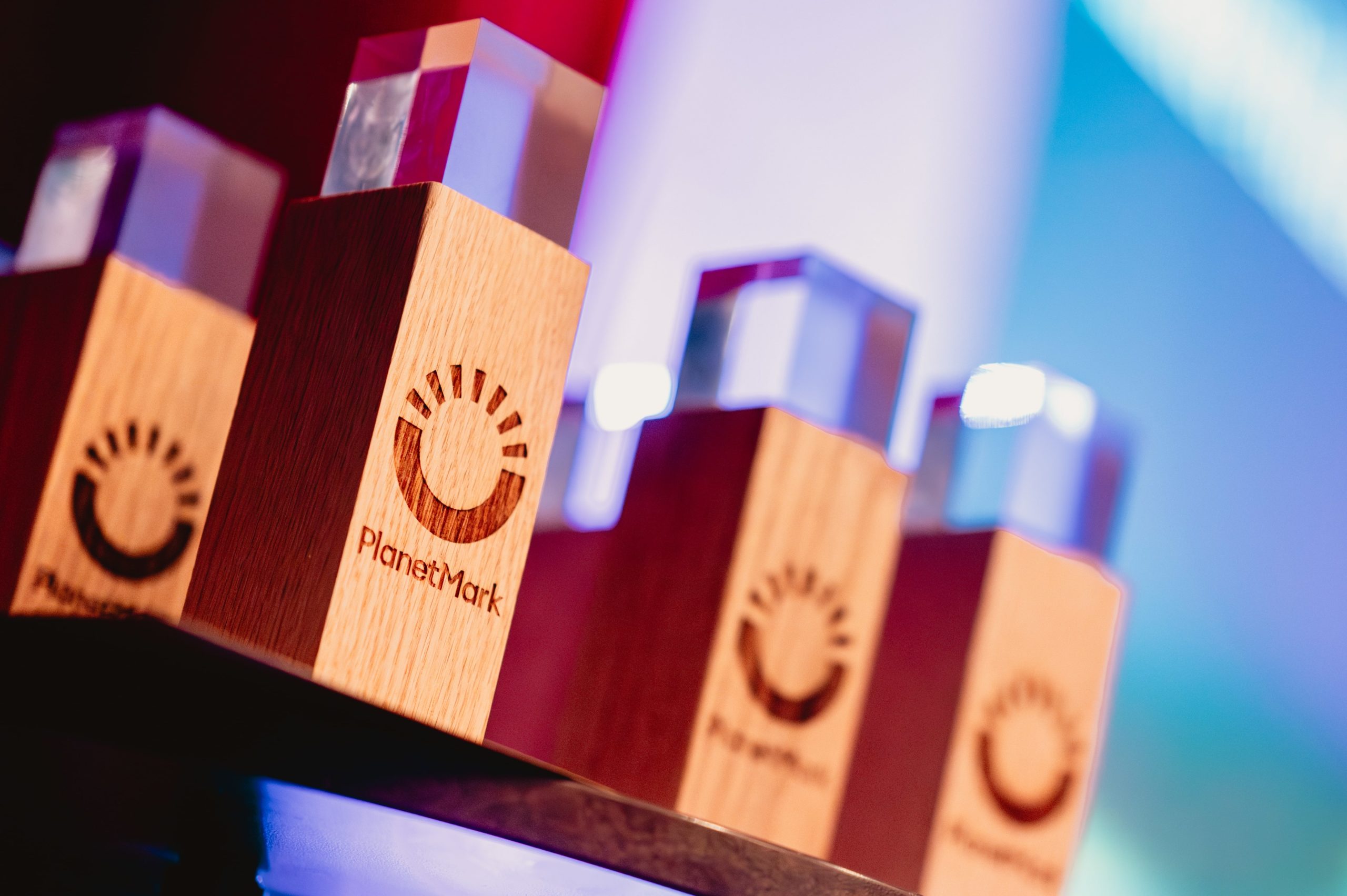 Four wooden trophies with clear square tops are displayed. Each trophy has a sunburst logo and the text planet mark engraved on the front. The background is softly blurred with blue, red, and white lighting.