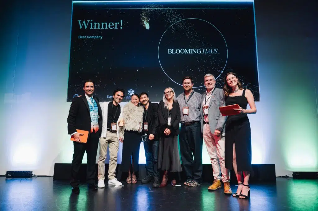 Eight people stand smiling on a stage under a large screen that reads "Winner! Best Company BLOOMING HAUS." Several hold orange objects, and all have conference lanyards. The background is dark with bright spotlights and celebratory graphics on the screen.