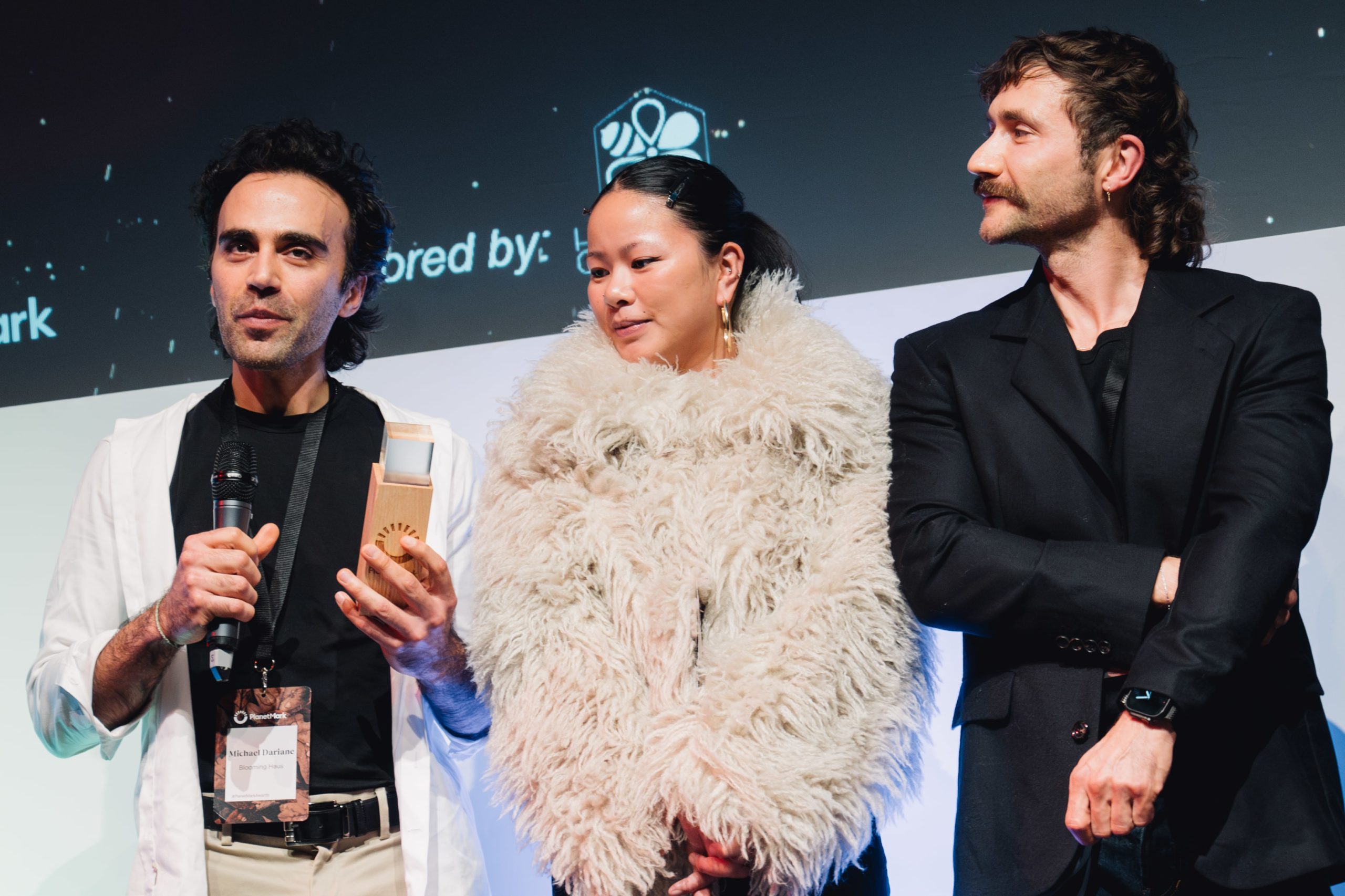 Three people stand on stage at the planet mark awards 2025. The man on the left holds a microphone and a trophy, wearing a white jacket. The woman in the centre wears a large cream-coloured coat. The man on the right stands in black with arms crossed.