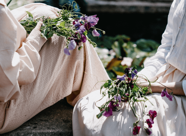 Two people in light dresses sitting outdoors, each holding a small bundle of sweet pea flowers