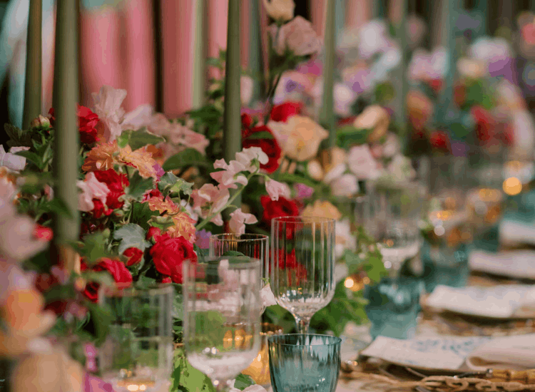 An elegant table setting with sweet pea arrangements in pink, purple, yellow, and red, surrounded by clear and blue glassware and tall green candles