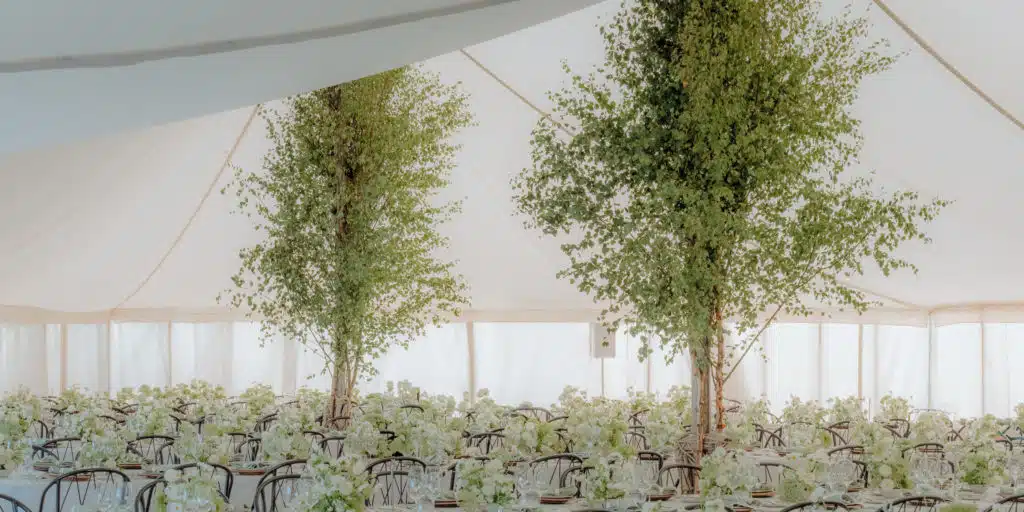 A white tent is set up for an event, with tall leafy trees placed inside among rows of tables. The tables are elegantly decorated by a luxury florist with white floral arrangements and glassware, creating a bright, airy, and sophisticated atmosphere.