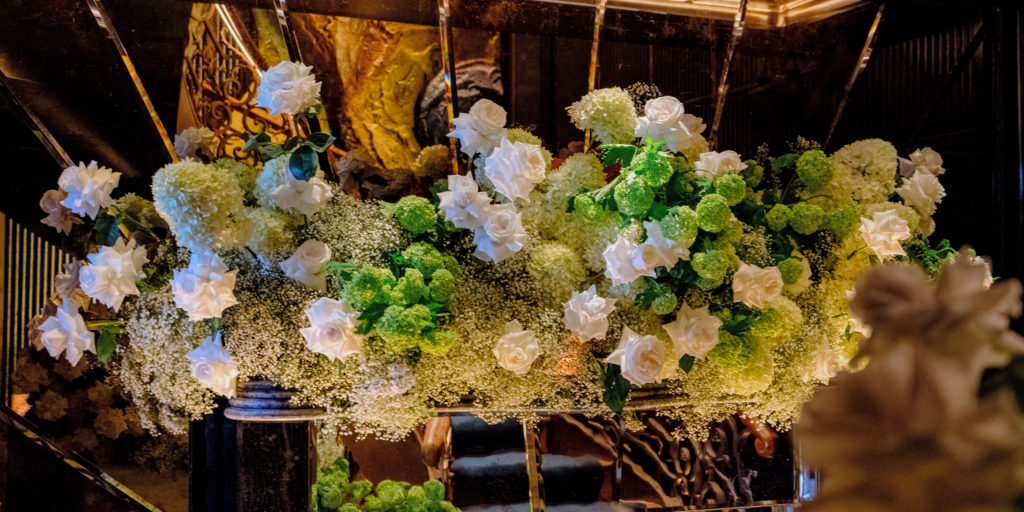 A lush floral arrangement by a luxury london florist features white roses, green hydrangeas, and baby's breath, displayed in a dark, elegant setting with ornate gold patterns and warm lighting, reflected in mirrors behind the bouquet.