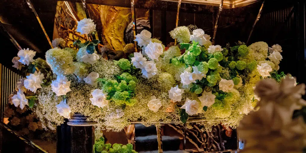 Corporate event flowers in london: what to expect 3 A lush floral arrangement by a luxury london florist features white roses, green hydrangeas, and baby's breath, displayed in a dark, elegant setting with ornate gold patterns and warm lighting, reflected in mirrors behind the bouquet.