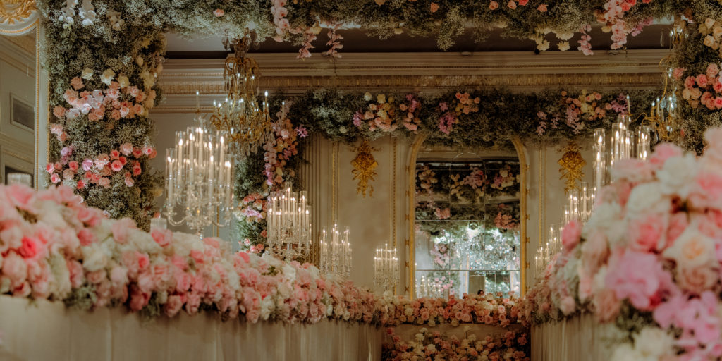 Opulent wedding aisle lined with pink and white blooms by a luxury florist, illuminated by crystal chandeliers. Floral arches and garlands adorn mirrored walls and ceiling, creating a romantic, luxurious setting with golden decorative details.