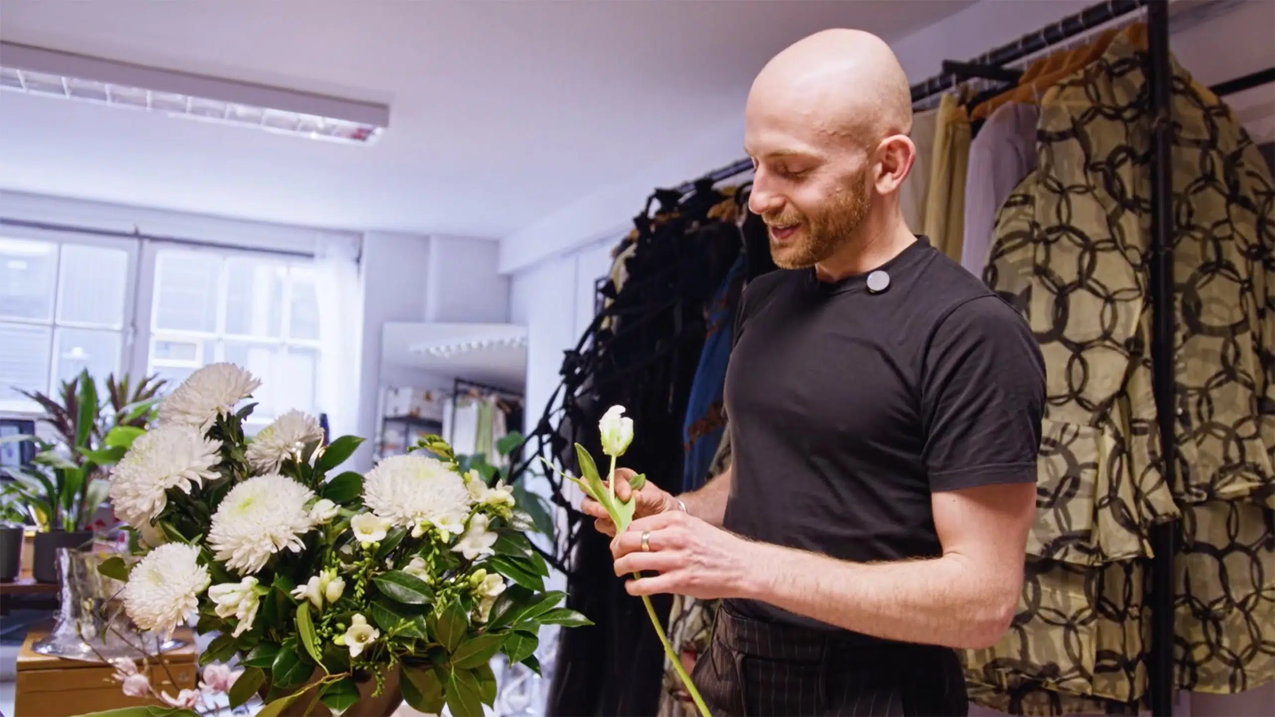 Patrick mcdowell's floral-themed fashion collections 4 Patrick mcdowell in a black t-shirt stands in a bright room, holding a white flower and arranging a bouquet of white flowers and greenery. Clothing racks with patterned garments are visible in the background.