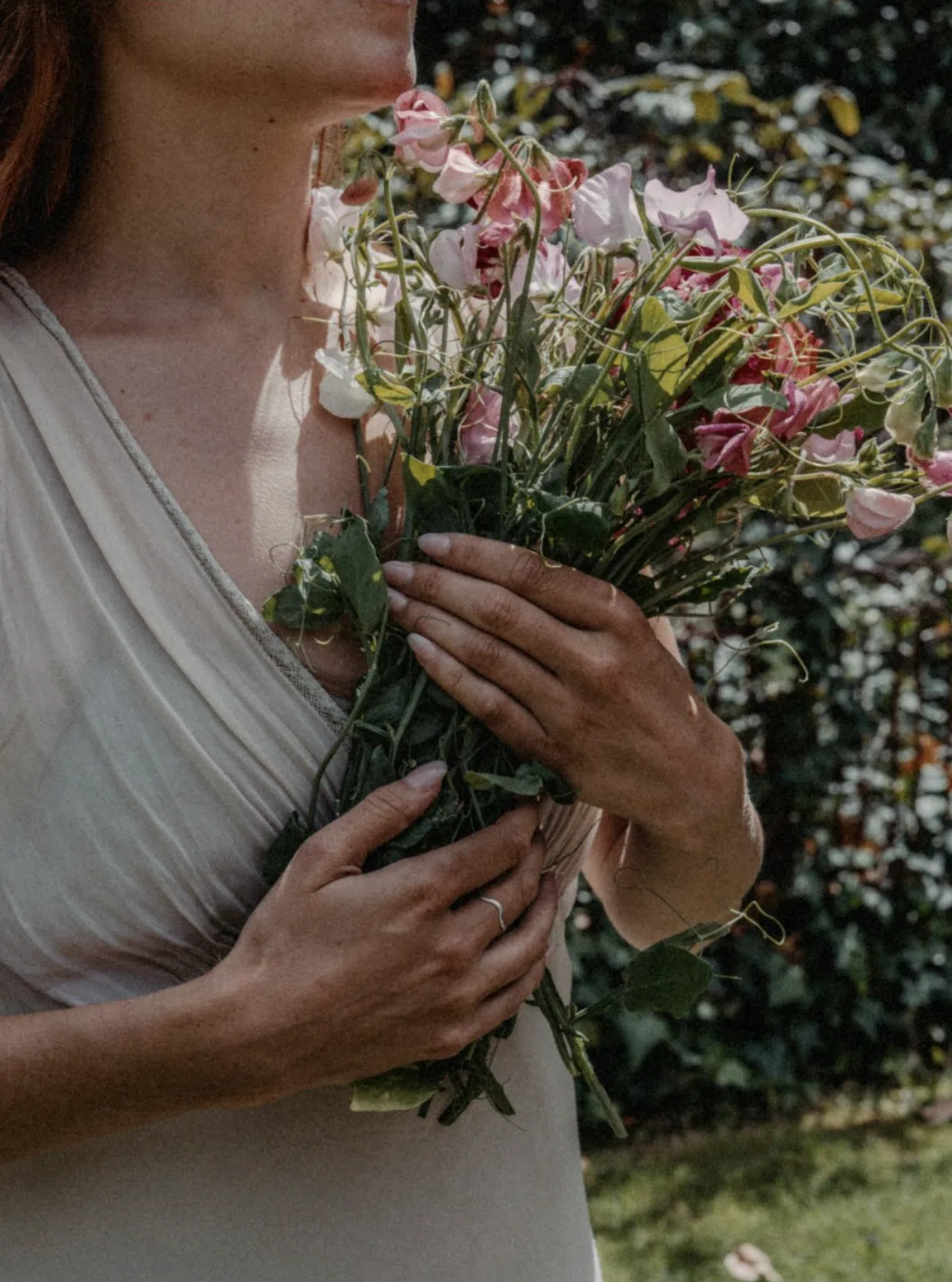 Sweet pea: blooming haus' flower of the year for 2026 18 A woman in a light sleeveless dress holding pink and magenta sweet pea flowers, standing outdoors with green foliage in the background