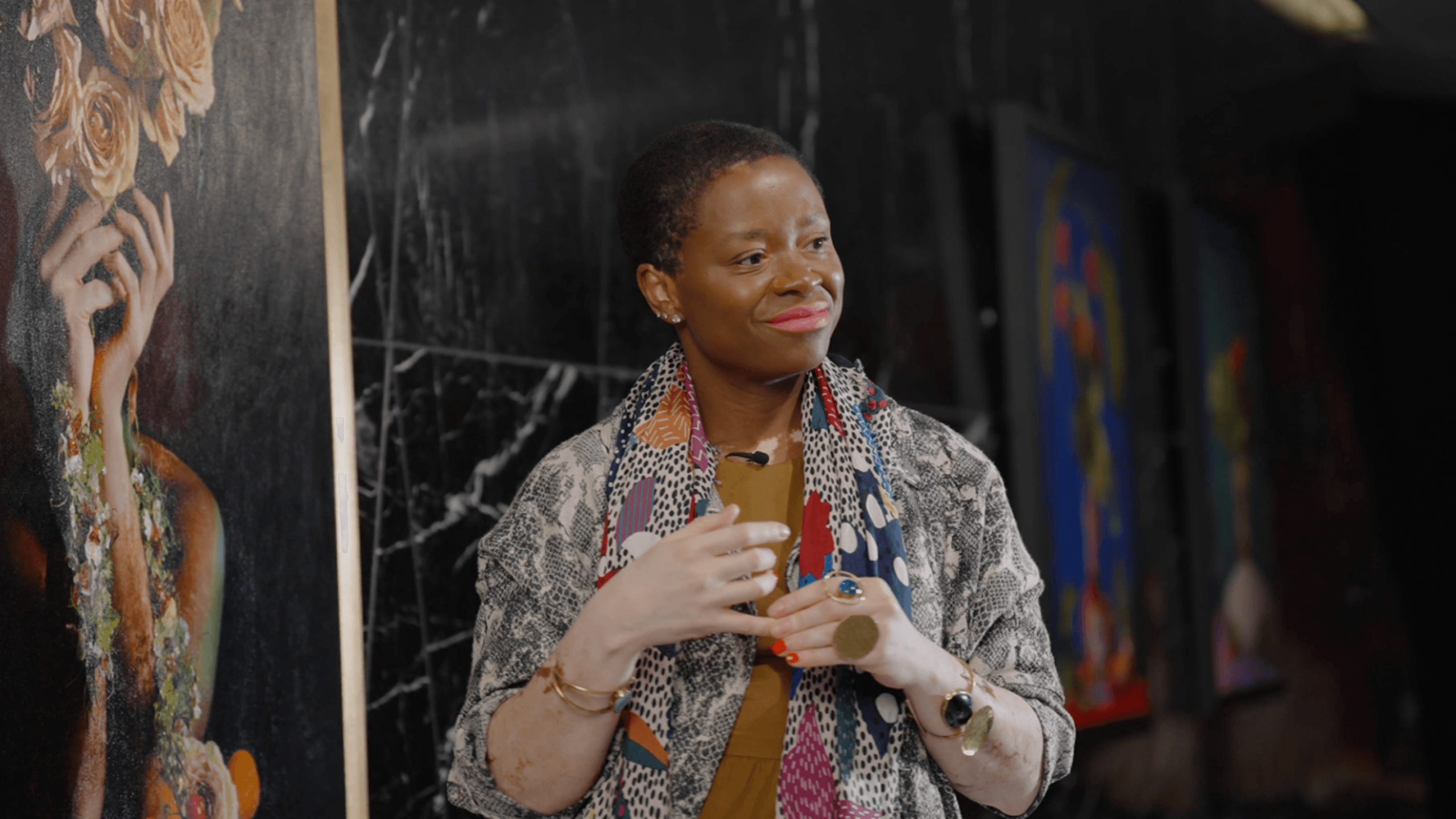 Violeta sofia indoors, wearing a colorful scarf, a patterned jacket, jewelry, and red lipstick. She smiles gently and clasps her hands together, with abstract art and a marble wall in the background.