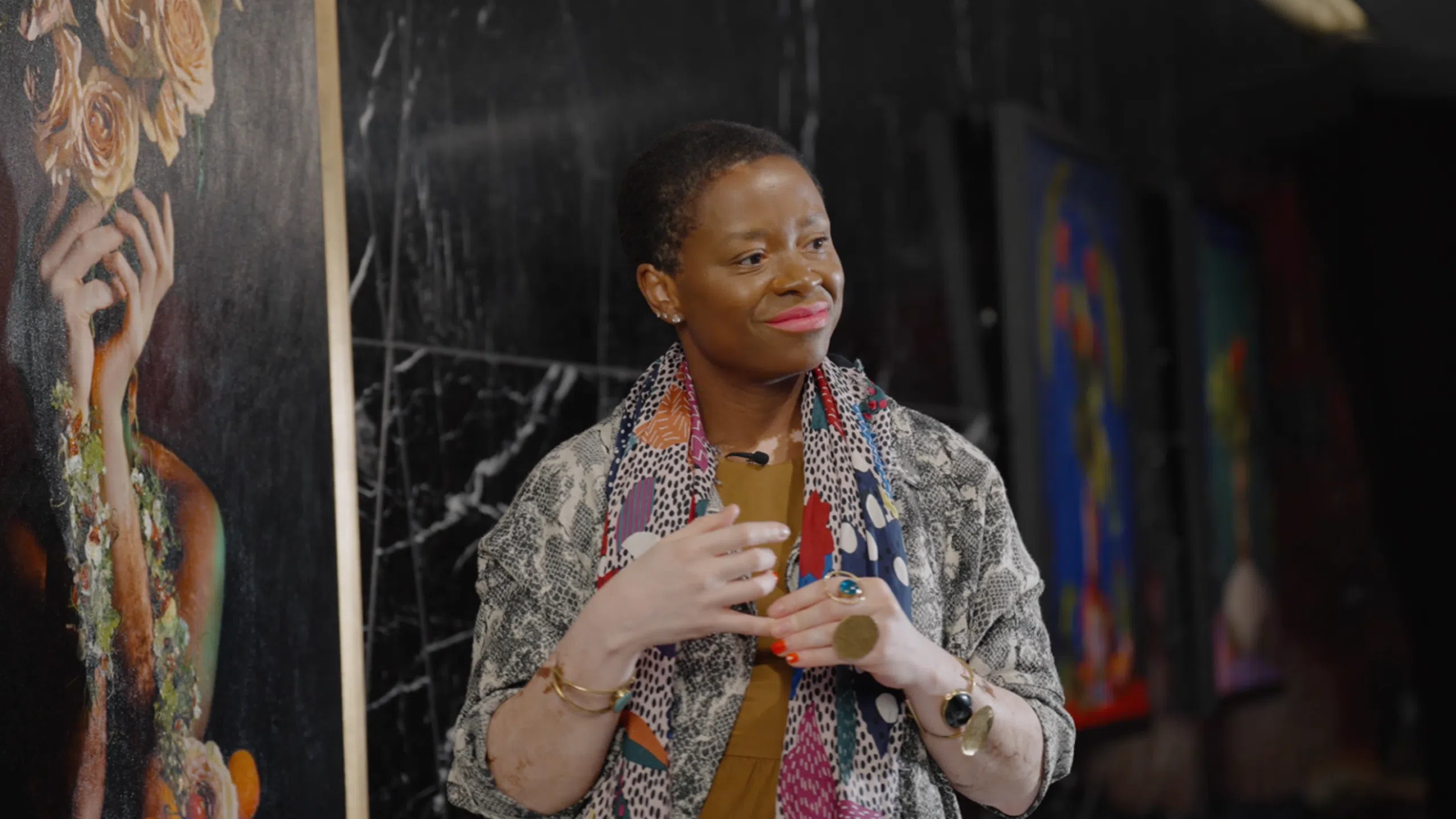 Artist violeta sofia's hand-masters series 2 Violeta sofia indoors, wearing a colorful scarf, a patterned jacket, jewelry, and red lipstick. She smiles gently and clasps her hands together, with abstract art and a marble wall in the background.
