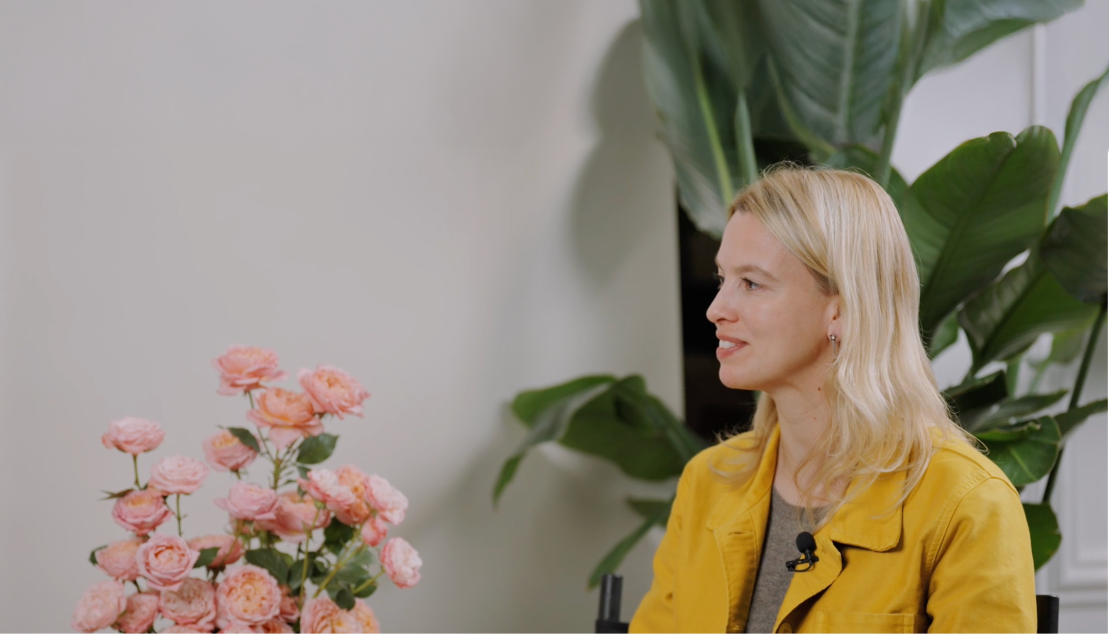Danae wearing a yellow jacket, sits indoors beside a vase of pink roses. She faces right, smiling, with large green plants and a blank light-coloured wall in the background. A small microphone is clipped to her jacket.